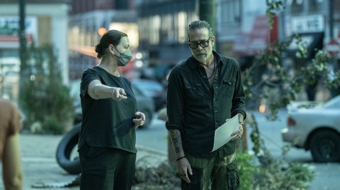 Behind-the-Scenes aus The Walking Dead: Dead City: Jeffrey Dean Morgan (Negan) steht mit einem Crewmitglied auf einer belebten Straße. Die Crewfrau trägt eine Maske und gibt Anweisungen, während Negan ein Skript in der Hand hält.