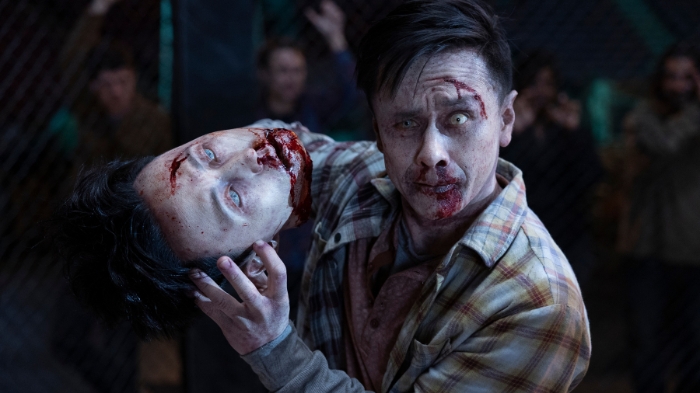Behind-the-Scenes aus The Walking Dead: Dead City: Ein als Zombie geschminkter Darsteller mit blutverschmiertem Gesicht hält eine realistische Requisite eines abgetrennten, identischen Kopfes. Im Hintergrund jubeln Zuschauer hinter einem Zaun.