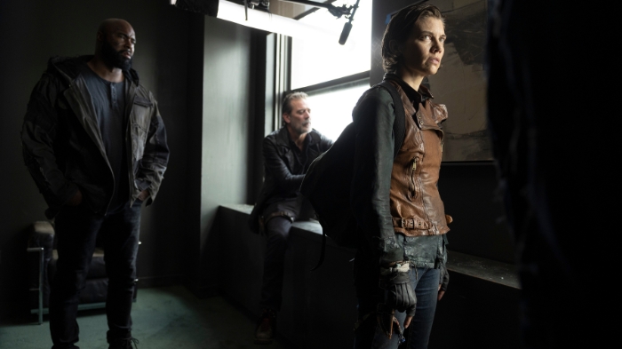 Behind-the-Scenes aus The Walking Dead: Dead City: Lauren Cohan (Maggie) steht mit ernster Miene in einem dunklen Raum, während Jeffrey Dean Morgan (Negan) am Fenster sitzt. Ein Crewmitglied hält eine Tonangel über die Szene.