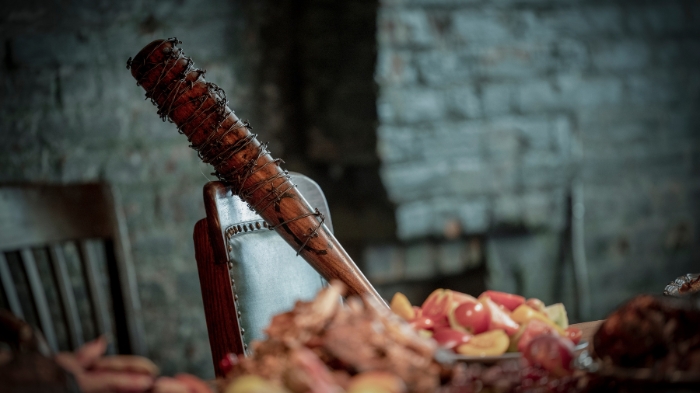 Behind-the-Scenes aus The Walking Dead: Dead City: Negans mit Stacheldraht umwickelter Baseballschläger Lucille lehnt an einem Stuhl inmitten eines üppigen Festmahls mit Fleisch und Obst.