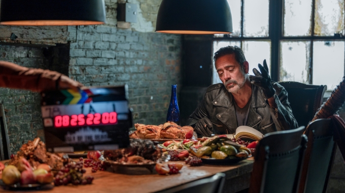 Behind-the-Scenes aus The Walking Dead: Dead City: Jeffrey Dean Morgan (Negan) sitzt an einem reich gedeckten Tisch mit Fleisch, Obst und Brot. Er trägt eine Lederjacke und hebt eine Hand, während eine Filmklappe die Szene markiert.