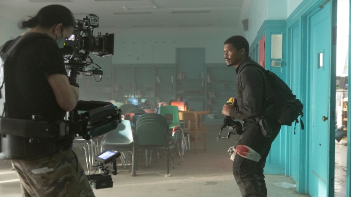 Behind-the-Scenes aus The Walking Dead: Dead City: Ein Kameramann filmt einen Darsteller mit Rucksack und Waffe, der angespannt in einem verwüsteten Klassenzimmer steht. Staub liegt in der Luft.