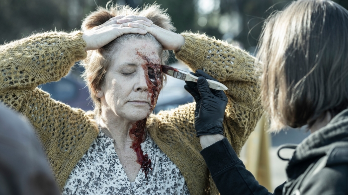 Behind-the-Scenes aus The Walking Dead: The Ones Who Live: Melissa McBride (Carol) mit geschlossenen Augen, während eine Maskenbildnerin ihr eine blutige Wunde ins Gesicht schminkt.