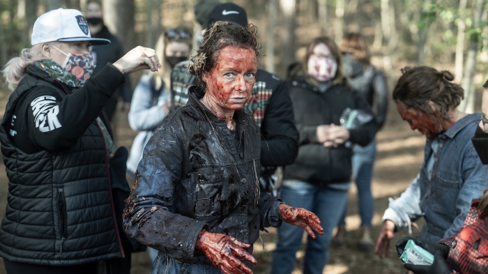 Behind-the-Scenes aus The Walking Dead: The Ones Who Live: Melissa McBride (Carol) steht blutverschmiert in dunkler Kleidung im Wald, während Maskenbildner und Crewmitglieder um sie herum arbeiten.