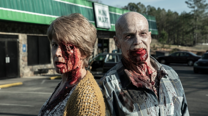 Behind-the-Scenes aus The Walking Dead: The Ones Who Live: Zwei als blutverschmierte Zombies geschminkte Darsteller stehen vor einem Supermarkt-Parkplatz und blicken in die Kamera.