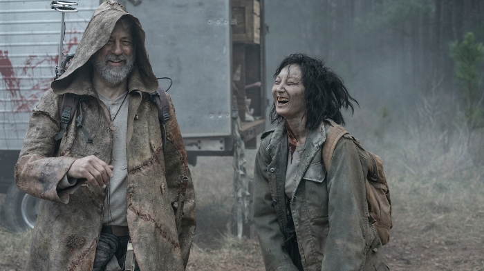 Behind-the-Scenes aus The Walking Dead: The Ones Who Live: Zwei Darsteller in abgenutzter, dreckiger Kleidung mit Rucksäcken lachen vor einem blutverschmierten Transporter in nebliger Waldkulisse.