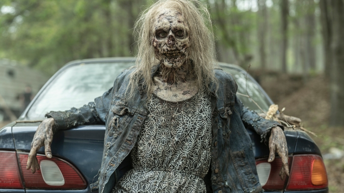Behind-the-Scenes aus The Walking Dead: The Ones Who Live: Eine als stark verweste Zombie-Frau geschminkte Darstellerin lehnt mit ausgebreiteten Armen an einem alten Auto im Wald.