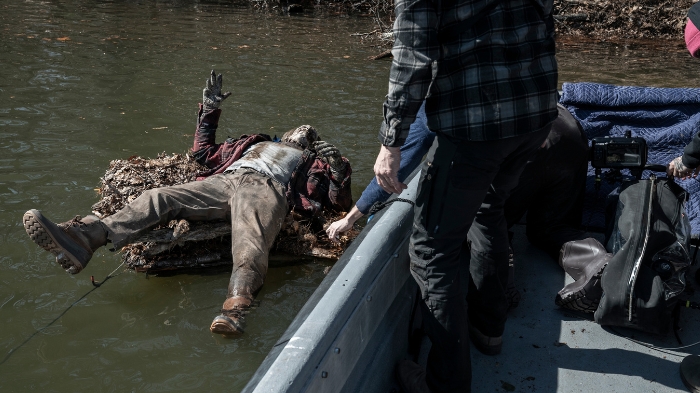 Behind-the-Scenes aus The Walking Dead: The Ones Who Live: Ein als Zombie geschminkter Darsteller treibt auf einer Plattform aus Ästen im Wasser, während Crewmitglieder aus einem Boot heraus an der Szene arbeiten