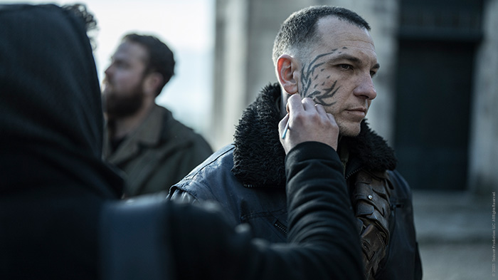 Ein Schauspieler wird von der Maskenbildnerin mit einem Tattoo im Gersicht versehen.
