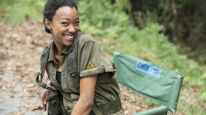 Behind-the-Scenes aus The Walking Dead: Sonequa Martin-Green als Sasha sitzt in ihrer grünen Klappstuhl mit Namensschild, trägt ihre Militärjacke und lächelt in die Kamera.