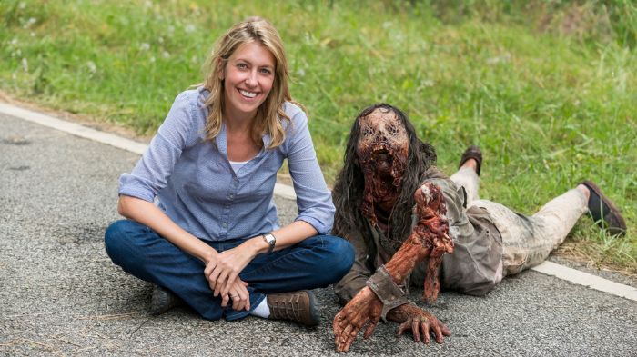 Behind-the-Scenes aus The Walking Dead: Eine Produzentin sitzt lächelnd auf der Straße neben einem detailliert geschminkten Zombie-Darsteller, der sich liegend abstützt.