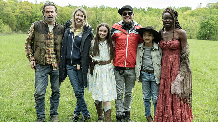 Behind-the-Scenes aus The Walking Dead: The Ones Who Live: Andrew Lincoln (Rick), Danai Gurira (Michonne), zwei junge Darstellerinnen, eine Crewmitglied-Frau und ein Regisseur posieren lachend auf einer Wiese.
