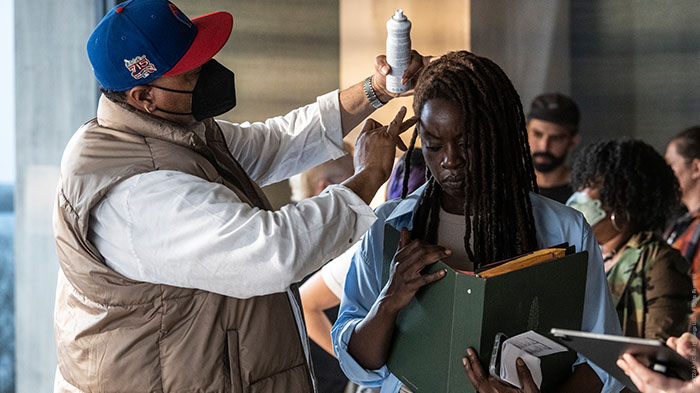 Behind-the-Scenes aus The Walking Dead: The Ones Who Live: Danai Gurira (Michonne) hält einen Aktenordner, während ein Crewmitglied ihr Haare und Make-up nachbessert. Weitere Crewmitglieder arbeiten im Hintergrund.