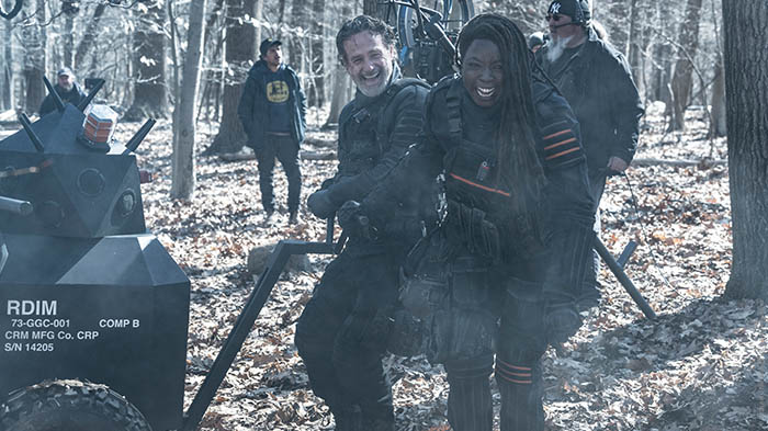 Behind-the-Scenes aus The Walking Dead: The Ones Who Live: Andrew Lincoln (Rick) und Danai Gurira (Michonne) lachen am Set in einem winterlichen Wald, umgeben von Crewmitgliedern und Kameraausrüstung.