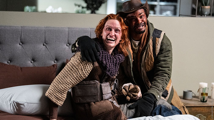 Behind-the-Scenes aus The Walking Dead: The Ones Who Live: Zwei Darsteller in postapokalyptischer Kleidung sitzen lachend auf einem Bett am Set, einer mit Hut und grünem Mantel, die andere mit rotem Haar und dicker Strickkleidung.