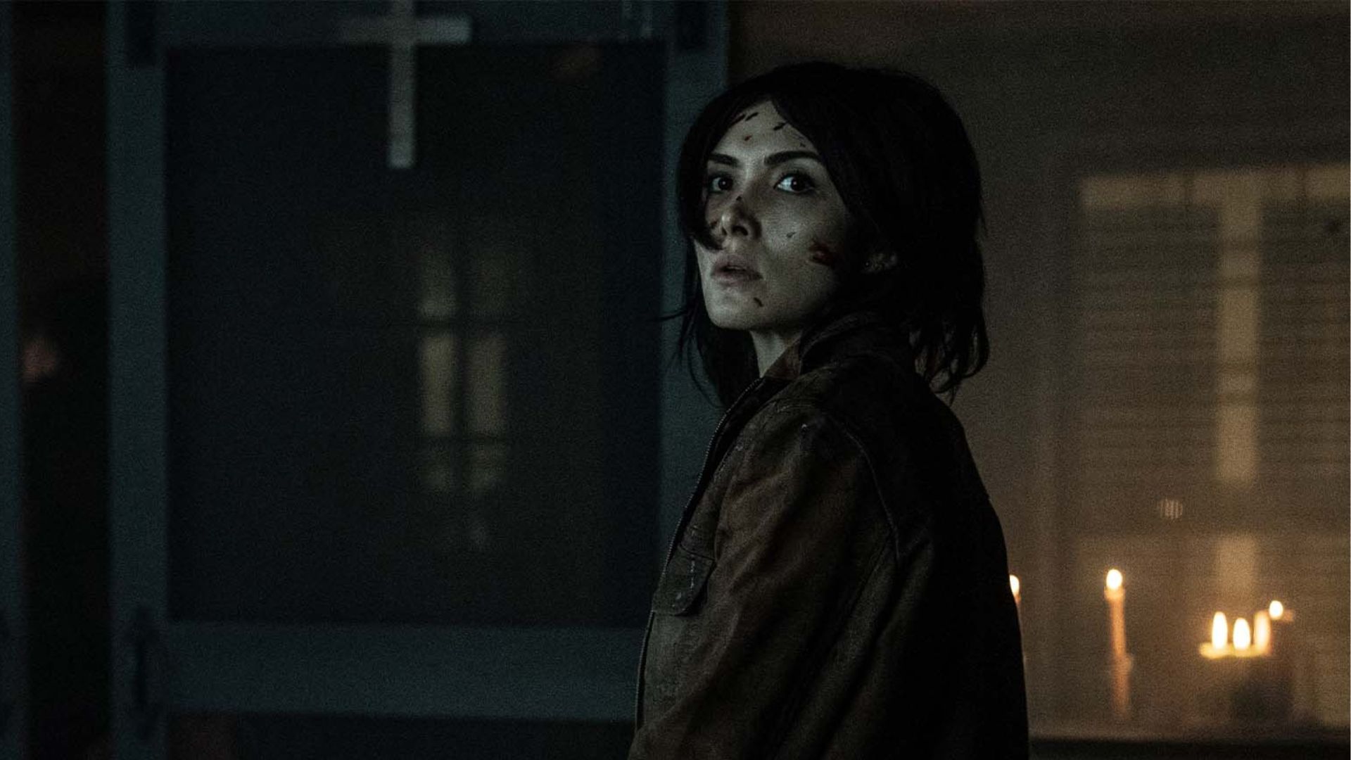Szenenbild aus The Walking Dead: The Ones Who Live: Eine junge Frau mit blutverschmiertem Gesicht und dunkler Jacke steht in einem düsteren Raum. Im Hintergrund brennen Kerzen, und ein Kreuz hängt an der Wand.